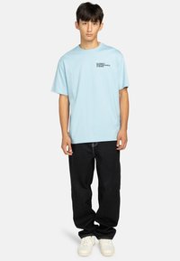 Camiseta de manga corta azul claro con pequeño texto negro en el pecho, combinada con unos vaqueros negros de corte ancho y zapatillas blancas.