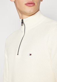 Detailní záběr osoby v texturované bílé mikině na čtvrt zip s malým červeným, bílým a modrým logem na hrudi.