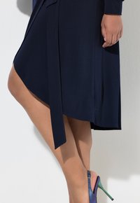 Navyblauwe wikkeljurk met een strik taille, met een diagonale zoom en een zachte, gladde textuur. Hoge hakken in iriserend blauw-groen.