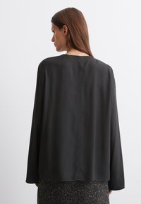 Black, long-sleeve blouse with a loose fit and round neckline, featuring smooth fabric and a clean back seam detail. Worn over a textured skirt.