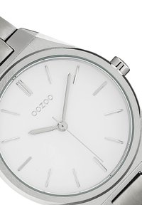 Montre en acier inoxydable argenté avec un cadran rond blanc, des marqueurs d'heure fins et une marque discrète. Elle présente un bracelet métallique lisse.