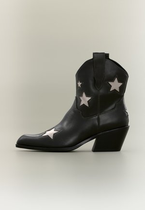 Bottine cowboy en cuir noir, hauteur cheville, avec un talon biseauté de hauteur moyenne et des découpes blanches en forme d’étoile sur les côtés et le bout.