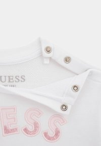 Valkoinen puuvillapaita, jossa on vaaleanpunaisista paljeteista tehty "GUESS"-logo. Kauluksessa olevat nepparit helpottavat pukemista. Pehmeä materiaali klassisella designilla.