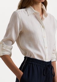 Camicia bianca in lino con colletto e maniche arrotolate, caratterizzata da bottoni. Indossata con pantaloni elasticizzati blu scuro in vita. Tessuto strutturato con una vestibilità casual.