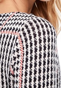 Pull en tricot avec un motif pied-de-poule noir et blanc agrémenté d'accents rouges, mettant en avant un tissu texturé et un design à épaules arrondies.