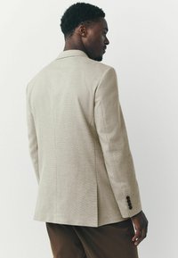 Blazer beige claro hecho de tela texturizada, con un corte entallado, solapas con muesca y tres botones negros en cada manga.