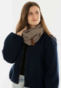 Gehaakte beige sjaal met een textuurpatroon, voorzien van een bruine leren label, gedragen over een donkere jas. Model poseert tegen een witte achtergrond.