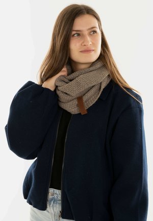 Gehaakte beige sjaal met een textuurpatroon, voorzien van een bruine leren label, gedragen over een donkere jas. Model poseert tegen een witte achtergrond.