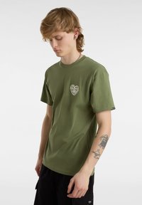 Camiseta de algodón verde oliva con un gráfico en forma de corazón que incluye el texto "VANS OF THE WALL". Cuello redondo, mangas cortas, corte relajado.