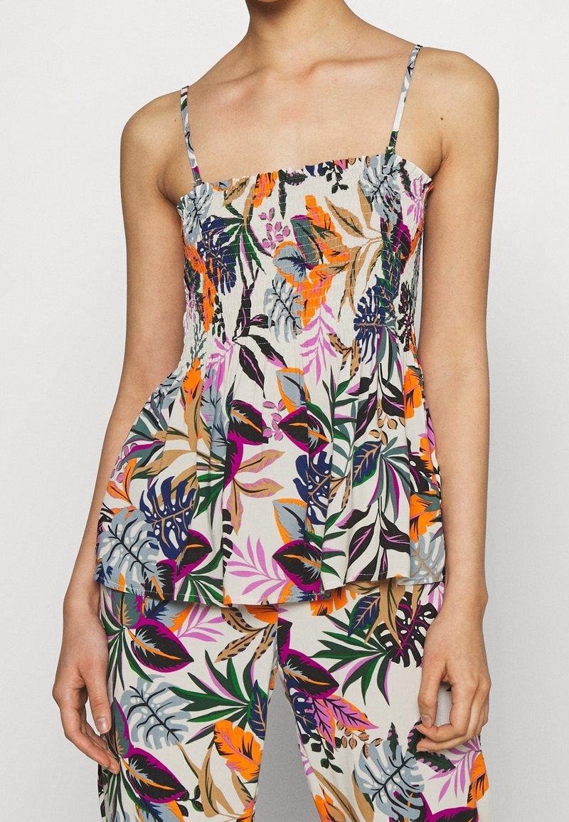 Femme portant un haut blanc à imprimé floral avec de fines bretelles et un pantalon assorti avec des feuilles tropicales orange, violettes, vertes et beiges.
