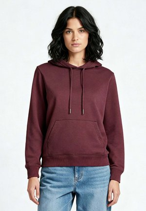 Donna con capelli neri ondulati che indossa una felpa con cappuccio color bordeaux con tasca frontale e jeans azzurri, in piedi contro uno sfondo chiaro e uniforme.