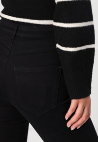 Zwarte high-waisted jeans met een gladde textuur, voorzien van versterkte stiknaden en een gebogen patch pocket. Wollen trui met witte strepen.