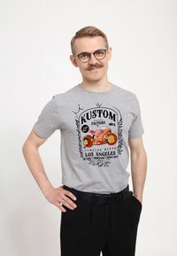 Grå bomulds t-shirt med en sort og farverig grafisk tryk med "KUSTOM FACTORY" og et motorcykel-design. Båret med sorte bukser.