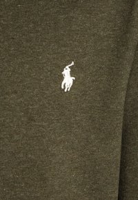 Camisa polo de algodón color verde oliva con un pequeño logo bordado en blanco de un caballo y un jinete en la parte izquierda del pecho. Textura suave.