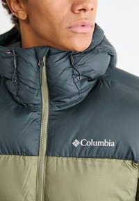 Jakna s kapuljačom koja ima gornji deo u tirkiznoj boji i donji deo u maslinasto zelenoj boji. Uključuje visoki ovratnik, zatvaranje na patent-zatvarač i Columbia logo na prsima.