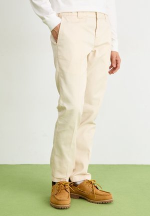 Beige chinos made of cotton with a straight-leg design, featuring side pockets and paired with brown shoes with yellow laces and textured soles.