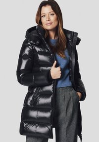 Czarna kurtka puchowa z kapturem, z pikowanymi sekcjami, wysokim kołnierzem i zamkiem błyskawicznym z przodu. Nosi się ją na jasnoniebieskim swetrze i teksturowanych szarych spodniach.