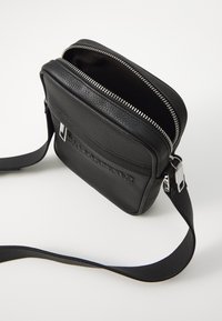 Černá kožená crossbody taška se zipovým uzávěrem, texturovaným povrchem, plochou přední kapsou, kovovými detaily v stříbrné barvě a nastavitelným popruhem.
