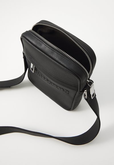 Sac en bandoulière en cuir noir avec fermeture éclair, surface texturée, poche avant plate, quincaillerie argentée et une bandoulière réglable.