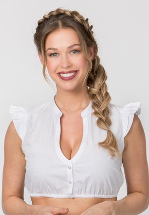 Lächelnde Frau mit geflochtenen Haaren trägt eine weiße, kurze Bluse und steht vor einem einfarbigen hellgrauen Hintergrund.