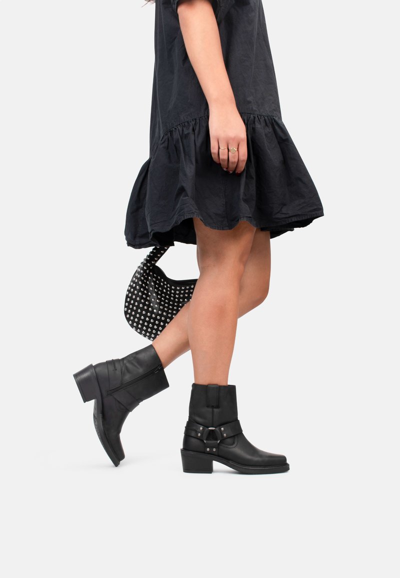 Bottines en cuir noir avec un petit talon carré, accent de boucle et détail de fermeture éclair, associées à une robe noire longueur genoux et un sac clouté.