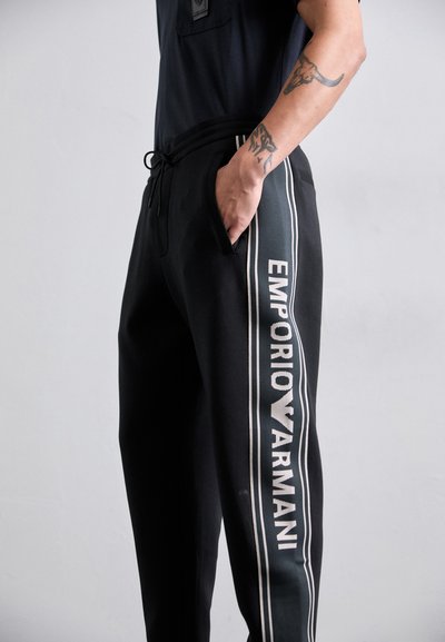 Mustaga Emporio Armani spordipüksid seljas isik, kellel on käsi taskus ja kaks tätoveeringut nähtavad küünarvarrel, hea kontrasti tagaplaanil.
