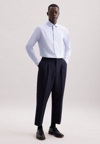 Camicia azzurra chiara con maniche lunghe, abbinata a pantaloni plissettati blu scuro e scarpe nere stringate. Tessuto morbido e vestibilità su misura.