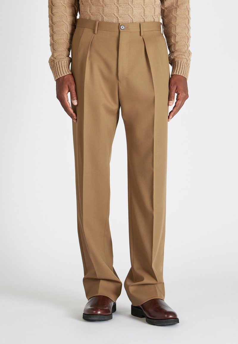 Beige gefaltete Hose mit taillierter Passform, aus einem glatten Stoff gefertigt. Mit Knopfverschluss und Taschen, kombiniert mit dunklen Schuhen.