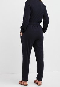 Jumpsuit de manga larga azul marino con cintura elástica, bolsillos y pantalones fluidos. Tela suave con corte relajado, combinado con sandalias planas.