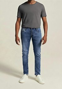 Man wearing gray short-sleeve T-shirt, blue slim-fit jeans, and white sneakers standing against a plain light background.