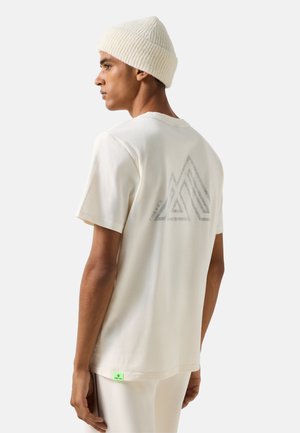 Jonge man met een witte gebreide muts en een gebroken wit T-shirt met een geometrisch ontwerp op de achterkant, zijwaarts kijkend op een effen achtergrond.