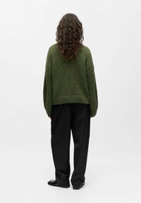 Grün, gestrickter Oversized-Pullover mit überschnittenen Schultern und geripptem Saum, kombiniert mit lockeren schwarzen Hosen und flachen schwarzen Schuhen.