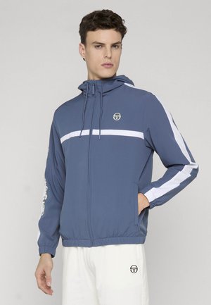 Giovane uomo che indossa una felpa con cappuccio blu con cerniera e strisce bianche, e pantaloni bianchi con un logo, in piedi contro uno sfondo chiaro e uniforme.