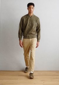 Olive green suede bomber jacket with ribbed cuffs, worn over light beige chinos and grey-brown sneakers. Model standing on wooden floor.