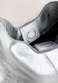 Ezüst színű bőr táska texturált felülettel. Lekerekített tetejű, patentgombbal záródó kialakítással és puha, tágas belső térrel rendelkezik.
