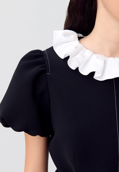 Vestido negro con mangas abullonadas, que presenta un cuello blanco con volantes y costuras contrastantes visibles. Textura de tela suave con un diseño ajustado.
