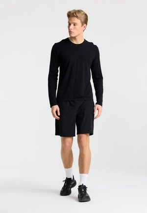 Ung mand iført sort langærmet skjorte, sorte shorts, hvide sokker og sorte sneakers står mod en ensartet lys baggrund.