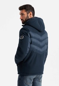 Marineblauer Puffer-Jacke mit Kapuze, die einen Chevron-Akzent in einer helleren Farbe, ein Logo-Patch an den Ärmeln und strukturierte Panels aufweist.