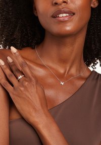 Collar de plata con un colgante de mariposa, acompañado de un anillo de plata con un diseño de mariposa; ambos están en la mano de un modelo de piel oscura.