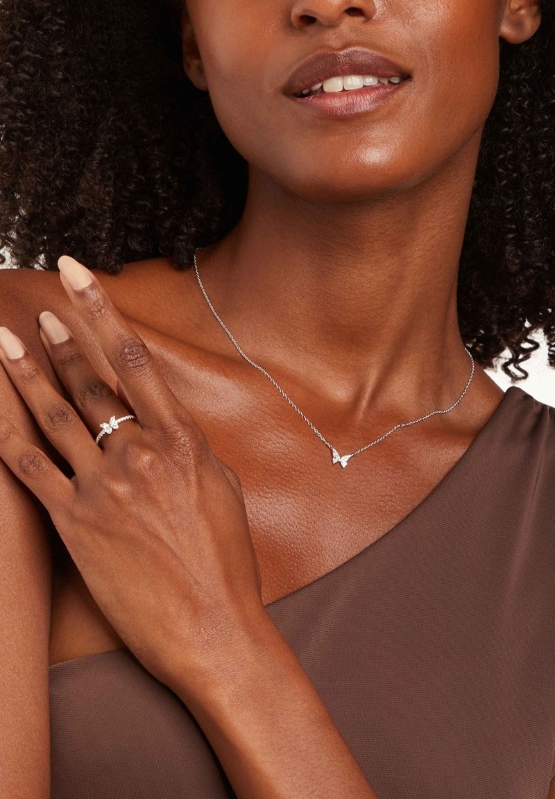 Collar de plata con un colgante de mariposa, acompañado de un anillo de plata con un diseño de mariposa; ambos están en la mano de un modelo de piel oscura.
