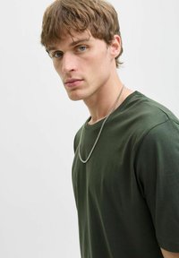 Camiseta de algodón verde oscuro con cuello redondo, combinada con un collar de cadena de plata. La tela parece suave y lisa, acompañada de una luz natural.