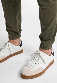 Hvide sneakers med tan ruskinds tå og brun gummisål. Har sort hæl accent og hvide snørebånd. Bæres med olivengrønne opfoldede bukser.