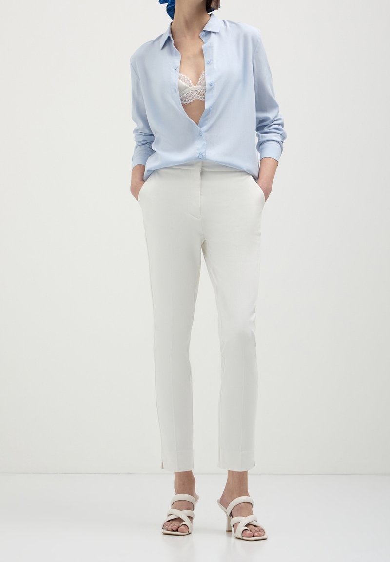 Femme portant une chemise bleu clair, un soutien-gorge blanc en dentelle, un pantalon blanc sur mesure et des sandales blanches à talons, debout les mains dans les poches.