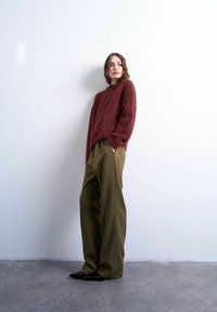 Femme aux cheveux ondulés portant un pull duveteux bordeaux, un pantalon large vert olive et des chaussures noires pointues, debout contre un mur blanc.