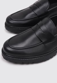Mocasines de cuero negro con una textura suave, detalle de correa en la parte delantera, punta redondeada y una suela gruesa y robusta para un mayor agarre.