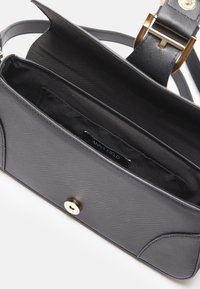Sac bandoulière noir texturé avec fermoir doré, bandoulière réglable et compartiment principal vide révélant l'étiquette de la marque à l'intérieur.