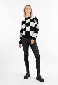 Schwarz-weißer Pullover mit Schachbrettmuster aus weichem, flauschigem Material; kombiniert mit schwarz glänzenden Lederähnlichen Hosen und klobigen schwarzen Stiefeln.