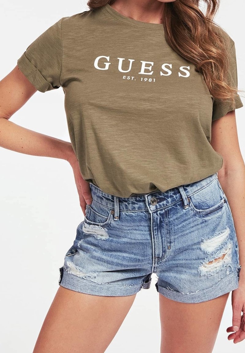 Olivengrøn bomulds t-shirt med hvid "GUESS" logo, parret med slidte denim shorts i lyseblå. Har rullede ærmer og frynsede kanter.