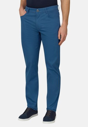 Mann trägt enganliegende blaue Hose, schwarzes Hemd und schwarze Turnschuhe mit weißen Sohlen, steht mit einer Hand in der Tasche und einer am Körper.