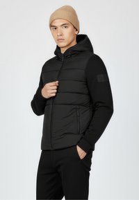 Black puffer jacket with a hood and ribbed cuffs; features smooth, shiny material, and side zip pockets. Worn with a beige knit beanie.